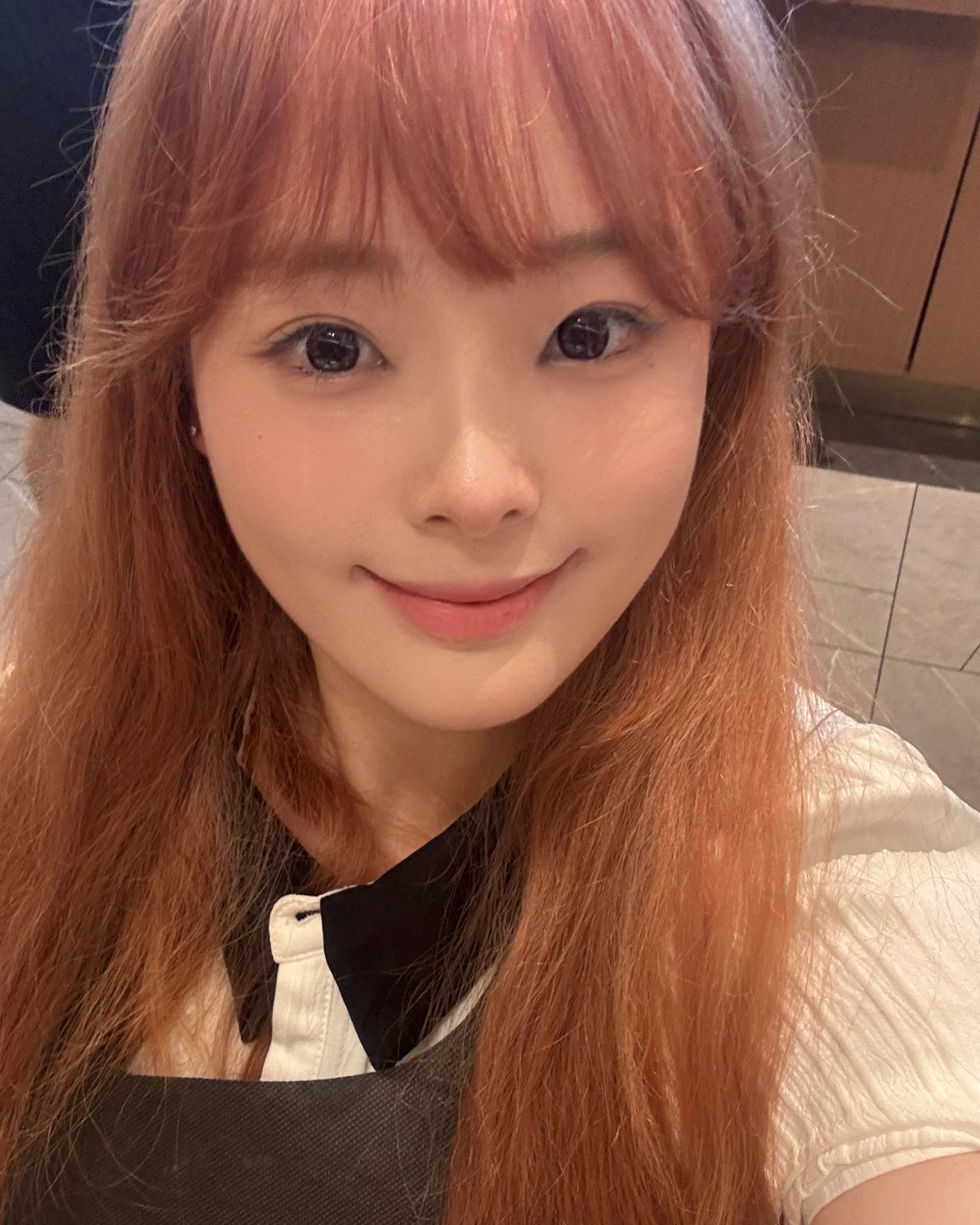 王宣懿 Diana 生活照：棕红色长发、齐刘海，微笑看向镜头，身穿白色上衣配深色背心裙，室内自拍